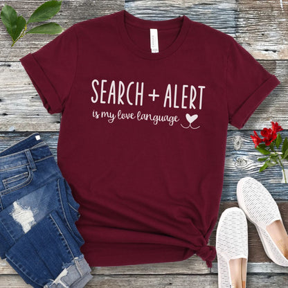 Maroon canine  scent work t-shirt with 'Search + Alert is my love language' text on a wooden surface with jeans and shoes.