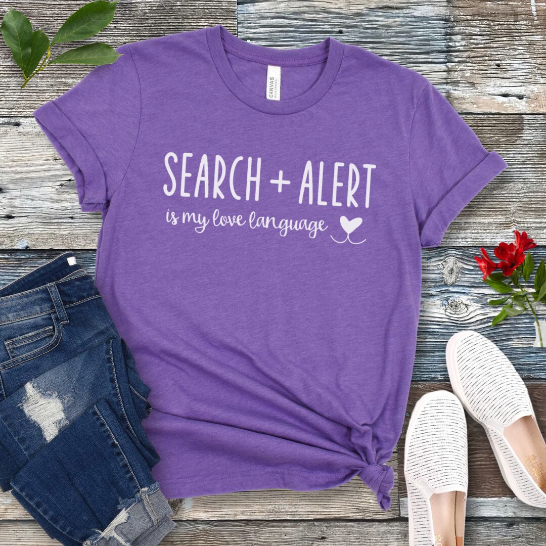 Purple dog scent training t-shirt with 'Search + Alert is my love language' text on a wooden surface with jeans and shoes.
