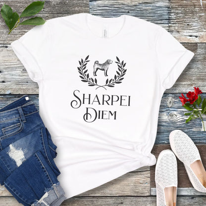 A white funny pun dog lover t-shirt with the text 'SHARPEI DIEM' and a graphic of a Shar Pei dog surrounded by laurel leaves. The shirt is displayed untied on a wooden surface with denim jeans, white shoes, and green leaves in the background.