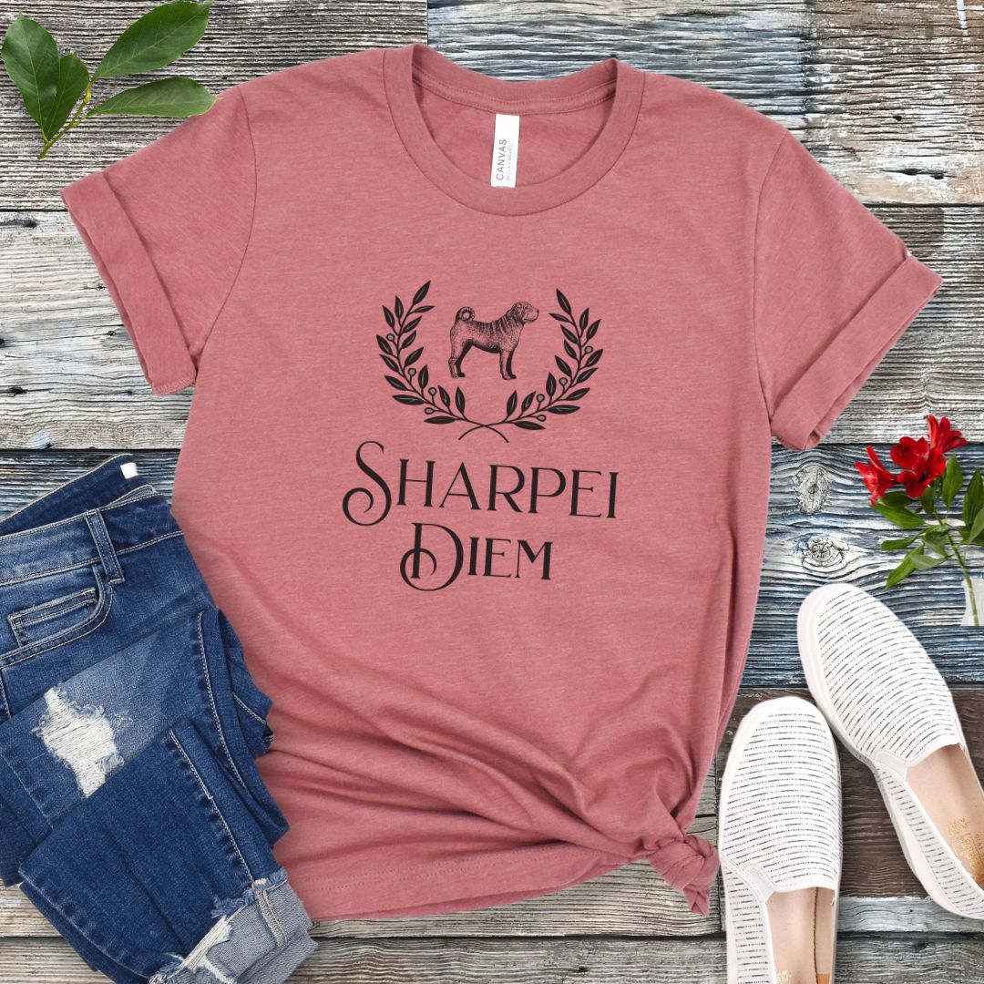 A mauve funny pun dog lover t-shirt with the text 'SHARPEI DIEM' and a graphic of a Shar Pei dog surrounded by laurel leaves. The shirt is displayed untied on a wooden surface with denim jeans, white shoes, and green leaves in the background.