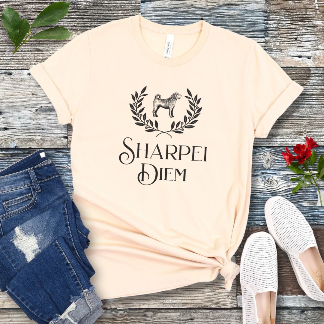 A beige funny pun dog lover t-shirt with the text 'SHARPEI DIEM' and a graphic of a Shar Pei dog surrounded by laurel leaves. The shirt is displayed untied on a wooden surface with denim jeans, white shoes, and green leaves in the background.