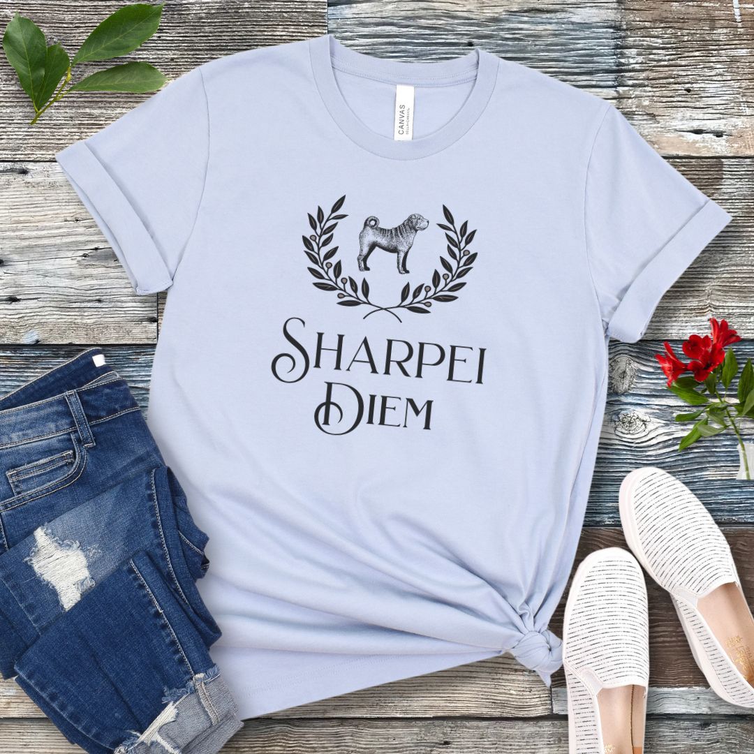 A light blue funny pun dog lover t-shirt with the text 'SHARPEI DIEM' and a graphic of a Shar Pei dog surrounded by laurel leaves. The shirt is displayed untied on a wooden surface with denim jeans, white shoes, and green leaves in the background.