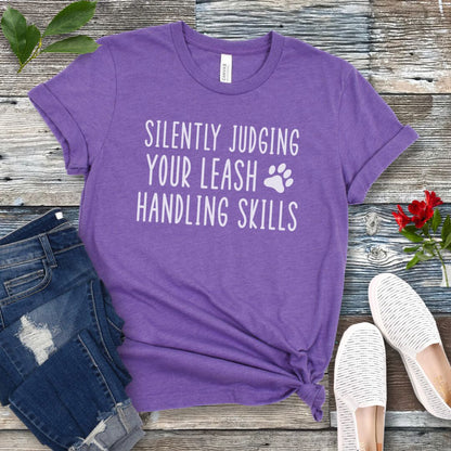 Purple unisex dog training t-shirt with humorous text, 'Silently Judging Your Leash Handling Skills' on a wooden surface with jeans and shoes.