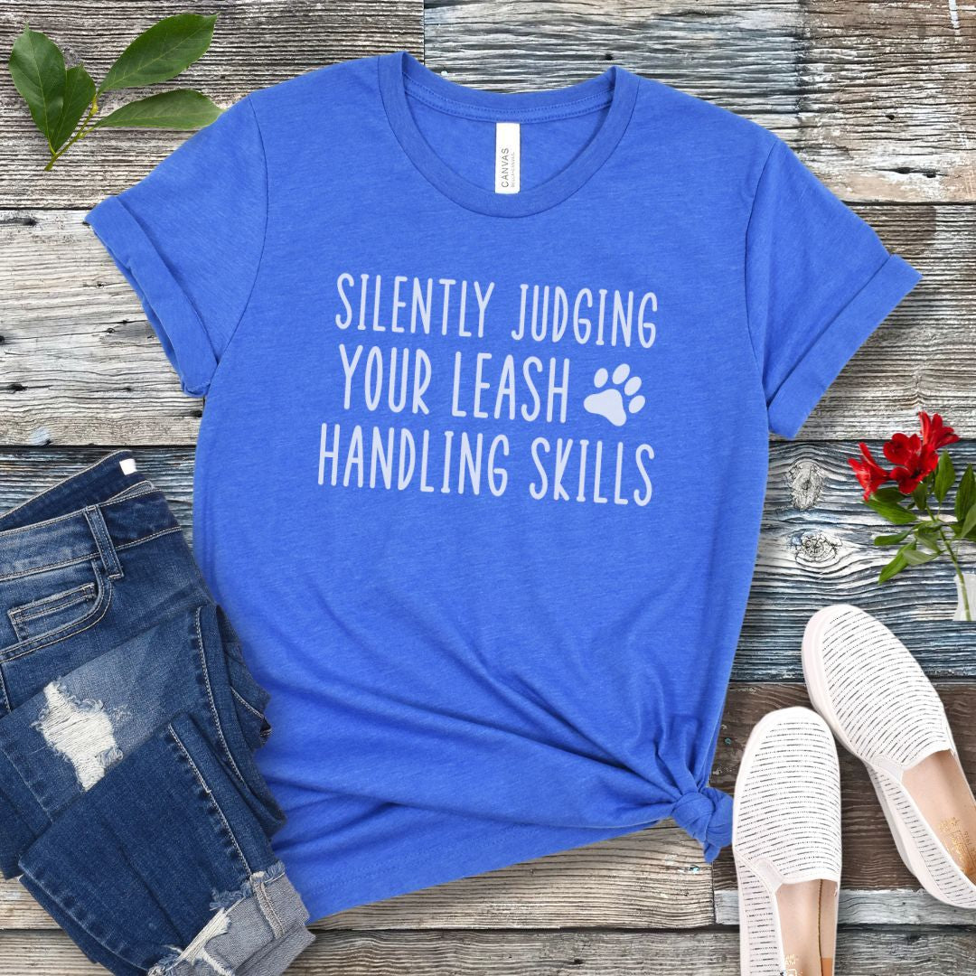 Royal blue unisex dog training t-shirt with humorous text, 'Silently Judging Your Leash Handling Skills' on a wooden surface with jeans and shoes.
