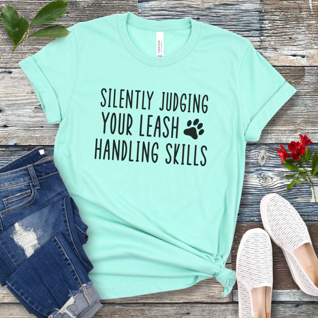 Mint green unisex dog training t-shirt with humorous text, 'Silently Judging Your Leash Handling Skills' on a wooden surface with jeans and shoes.