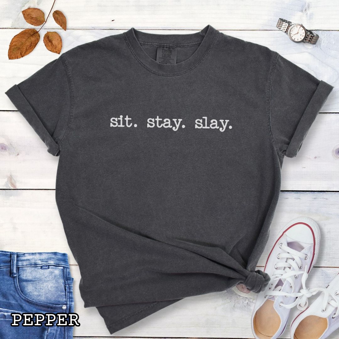 Grey (Pepper) Comfort Colors dog training t-shirt with 'sit. stay. slay.' text on a wooden surface with jeans, sneakers, and sunglasses.