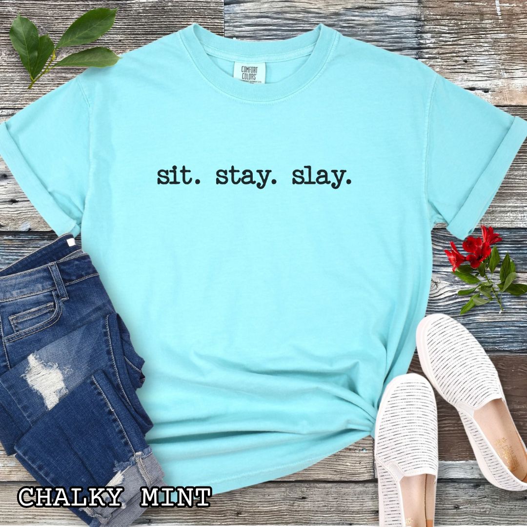 Light teal (Chalky Mint) Comfort Colors dog training t-shirt with 'sit. stay. slay.' text on a wooden surface with jeans, sneakers, and sunglasses.