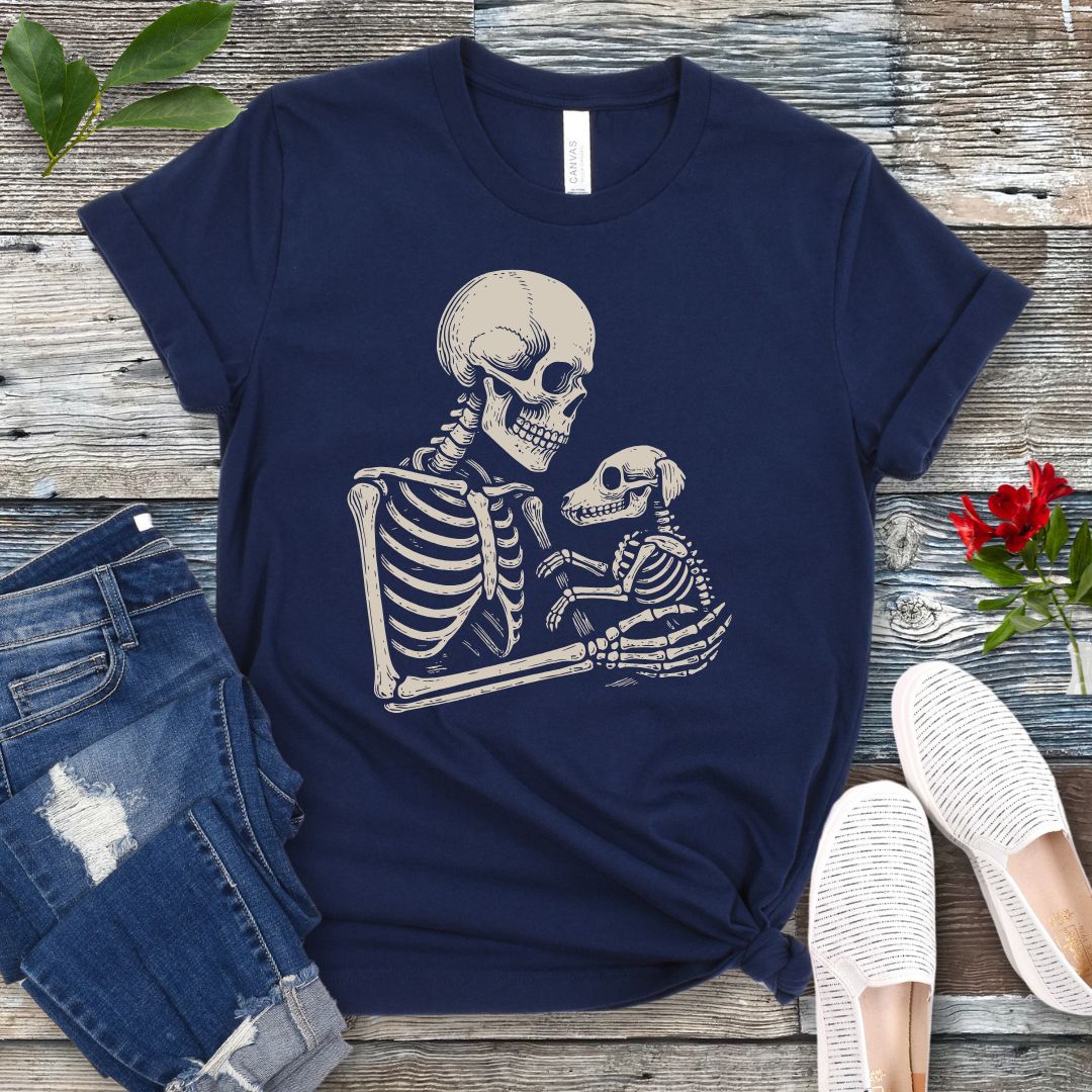 A navy blue t-shirt for dog lovers with a monochrome vintage-style illustration of a skeleton holding a skeletal dog, displayed on a wooden surface with denim jeans and white shoes in the background.