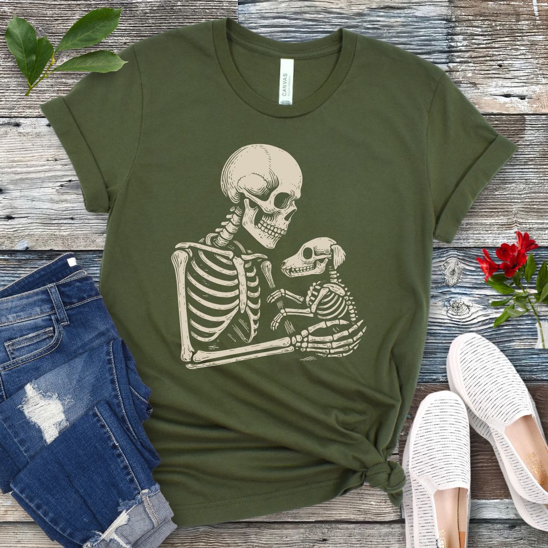 A military green t-shirt for dog lovers with a monochrome vintage-style illustration of a skeleton holding a skeletal dog, displayed on a wooden surface with denim jeans and white shoes in the background.