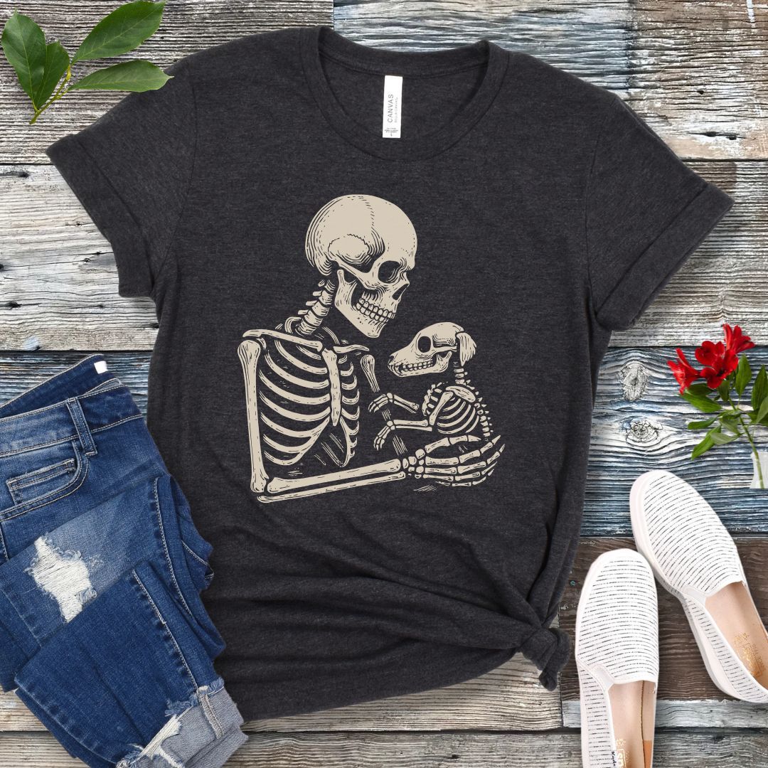 A dark grey t-shirt for dog lovers with a monochrome vintage-style illustration of a skeleton holding a skeletal dog, displayed on a wooden surface with denim jeans and white shoes in the background.