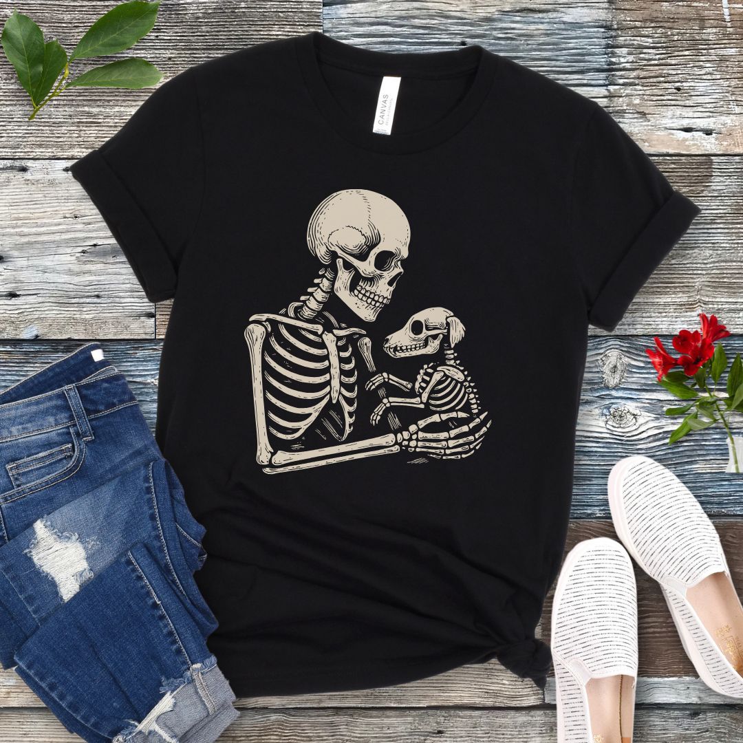 A black t-shirt for dog lovers with a monochrome vintage-style illustration of a skeleton holding a skeletal dog, displayed on a wooden surface with denim jeans and white shoes in the background.