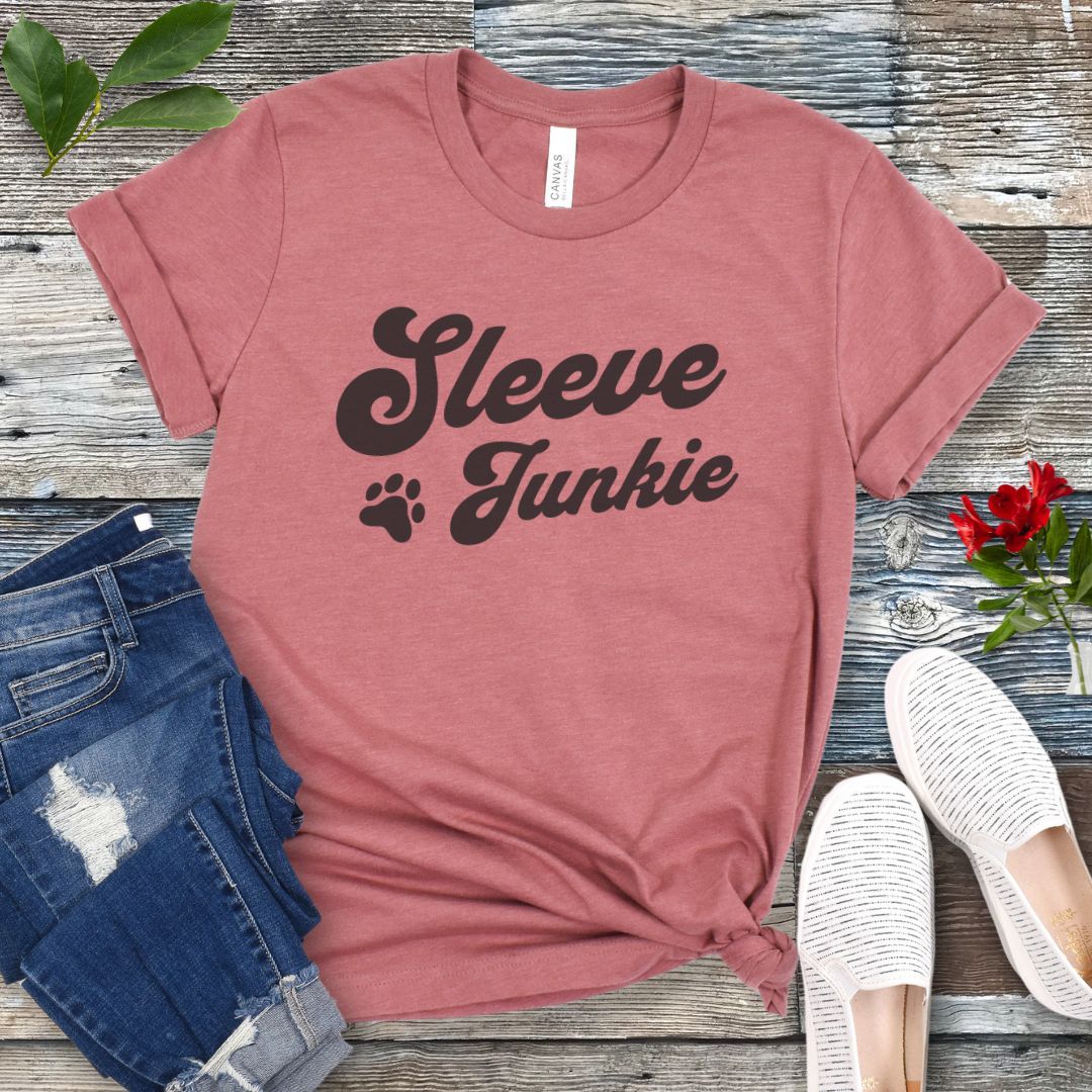 Mauve IGP bite work t-shirt with 'Sleeve Junkie' text bold, retro font and paw print on a wooden surface with jeans and shoes.