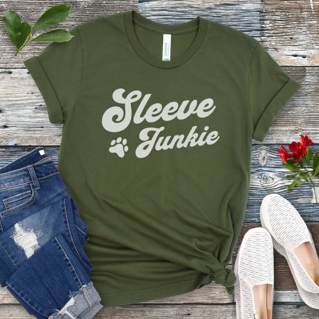 Military green IGP bite work t-shirt with 'Sleeve Junkie' text bold, retro font and paw print on a wooden surface with jeans and shoes.