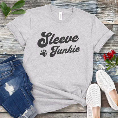 Light grey IGP bite work t-shirt with 'Sleeve Junkie' text bold, retro font and paw print on a wooden surface with jeans and shoes.