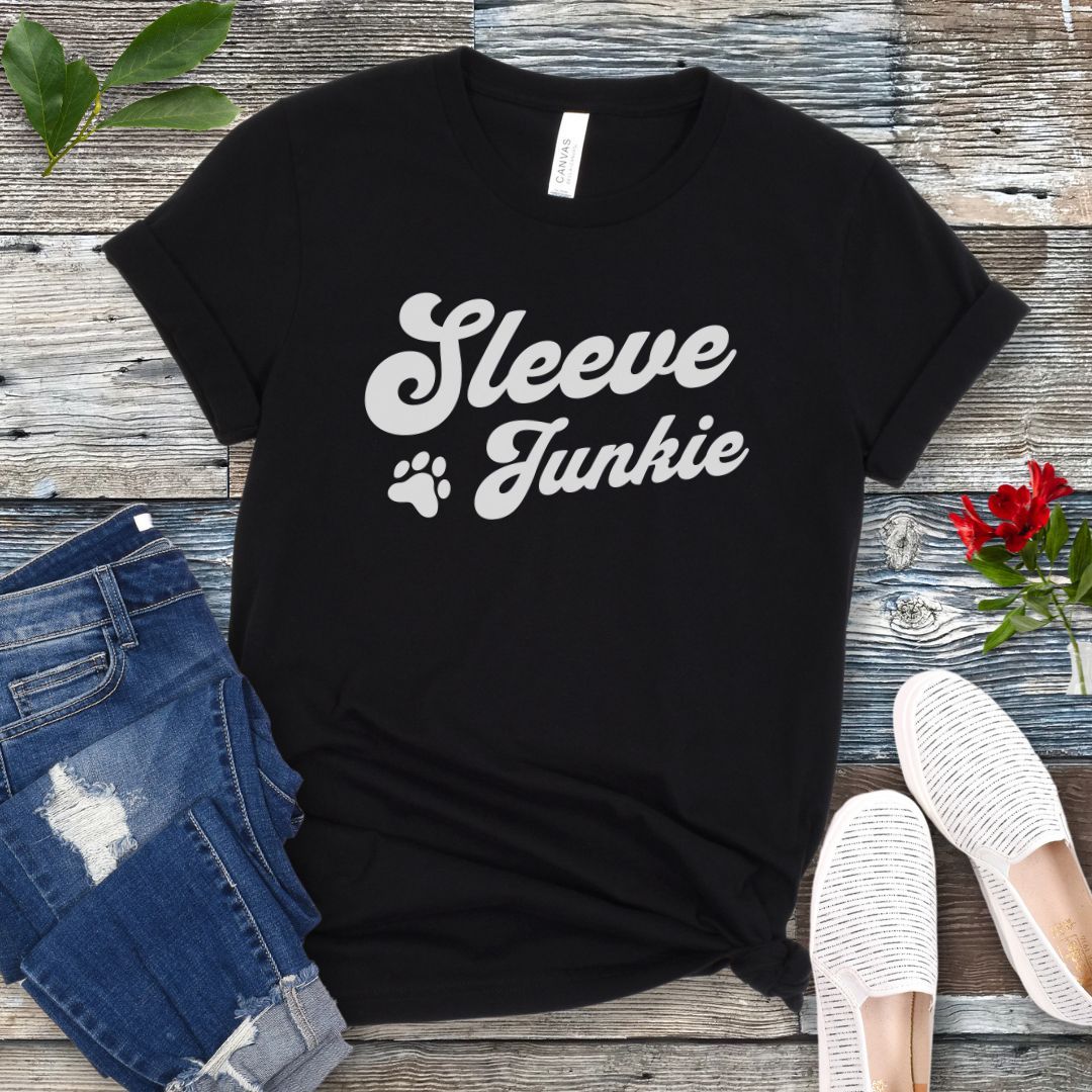 Black IGP bite work t-shirt with 'Sleeve Junkie' text bold, retro font and paw print on a wooden surface with jeans and shoes.