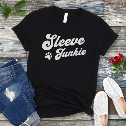 Black IGP bite work t-shirt with 'Sleeve Junkie' text bold, retro font and paw print on a wooden surface with jeans and shoes.