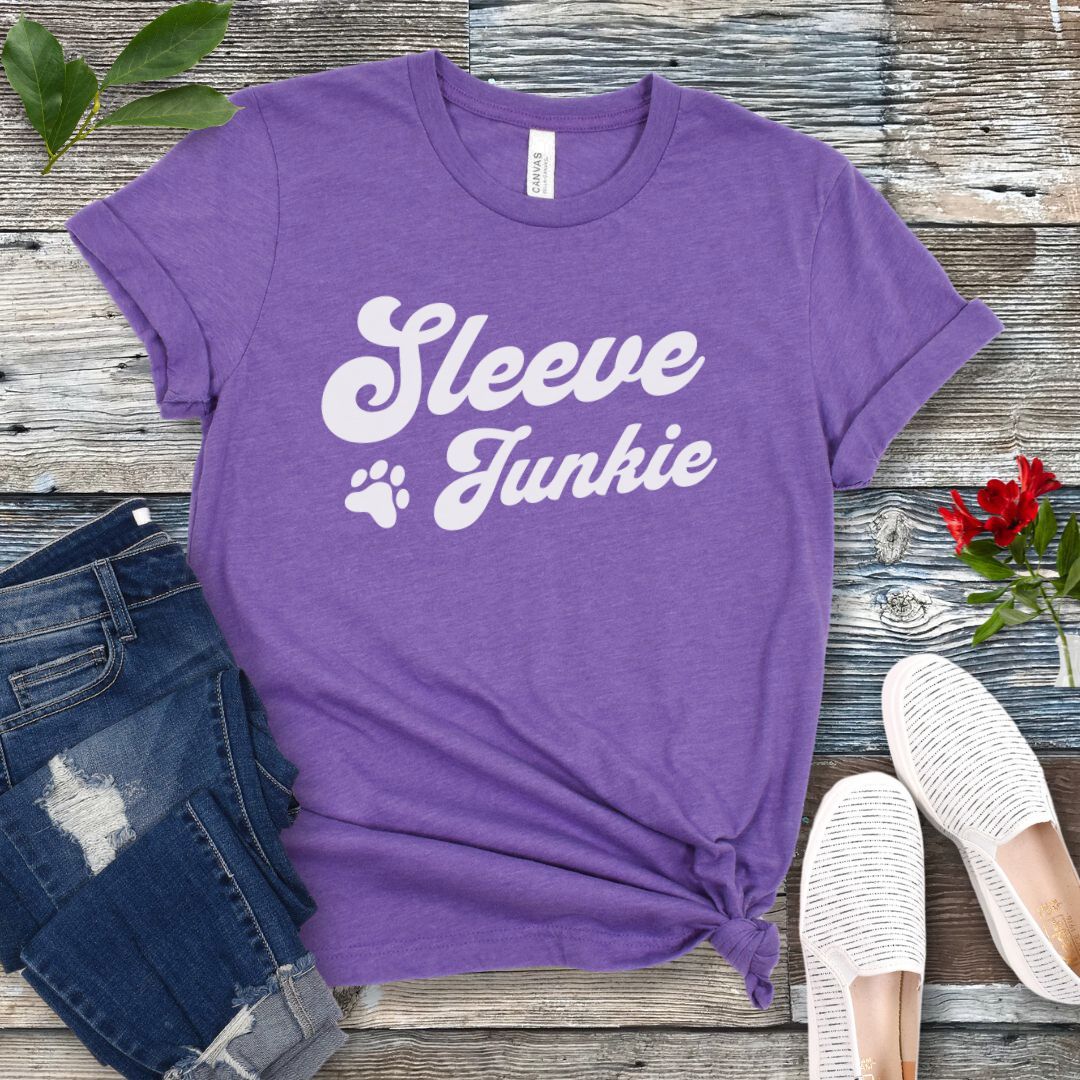Purple IGP bite work t-shirt with 'Sleeve Junkie' text bold, retro font and paw print on a wooden surface with jeans and shoes.