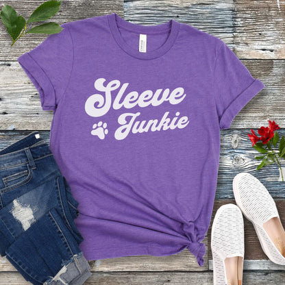 Purple IGP bite work t-shirt with 'Sleeve Junkie' text bold, retro font and paw print on a wooden surface with jeans and shoes.