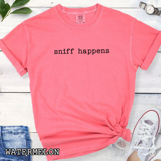 Pink Comfort Colors nosework t-shirt with 'sniff happens' text on a wooden surface with jeans and sneakers.