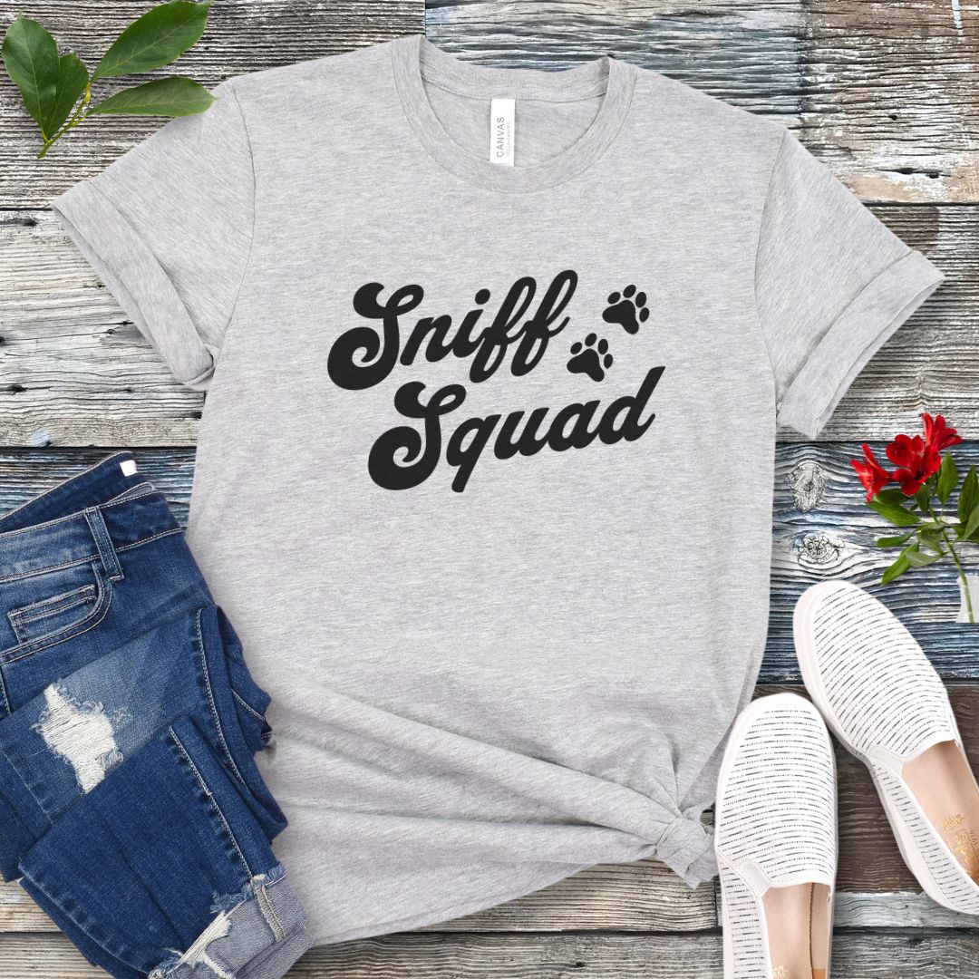 Light grey canine scent work t-shirt with 'Sniff Squad' text in retro font and paw prints on a wooden surface with jeans and shoes.