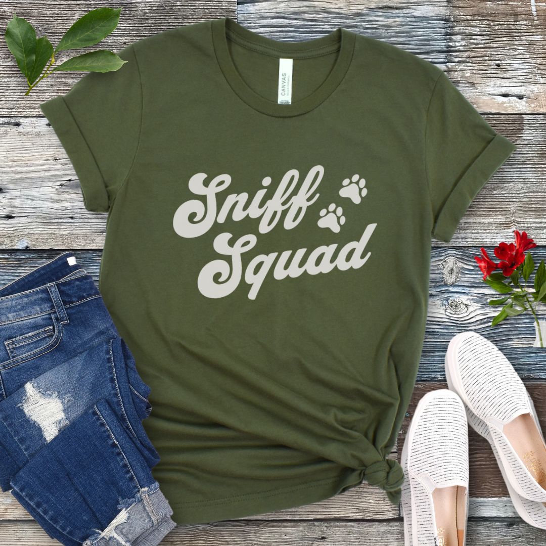 Military green K9 nose work t-shirt with 'Sniff Squad' text in retro font and paw prints on a wooden surface with jeans and shoes.