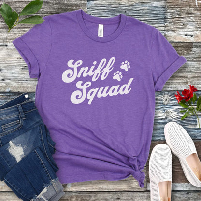 Purple canine scent work t-shirt with 'Sniff Squad' text in retro font and paw prints on a wooden surface with jeans and shoes.