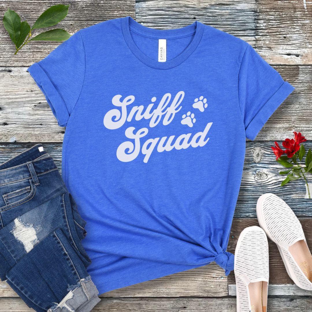 Royal blue canine scent work t-shirt with 'Sniff Squad' text in retro font and paw prints on a wooden surface with jeans and shoes.