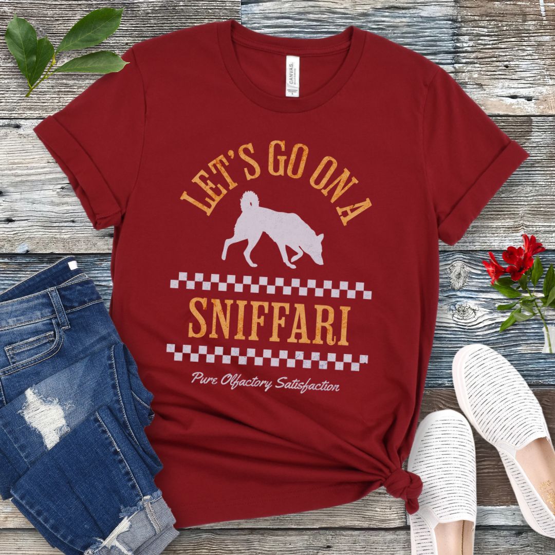 A dark red t-shirt with 'LET'S GO ON A SNIFFARI' text and a silhouette of a sniffing dog in the center, displayed flat with a pair of blue jeans and white shoes on a wooden surface.