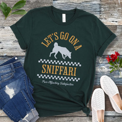A forest green t-shirt with 'LET'S GO ON A SNIFFARI' text and a silhouette of a sniffing dog in the center, displayed flat with a pair of blue jeans and white shoes on a wooden surface.