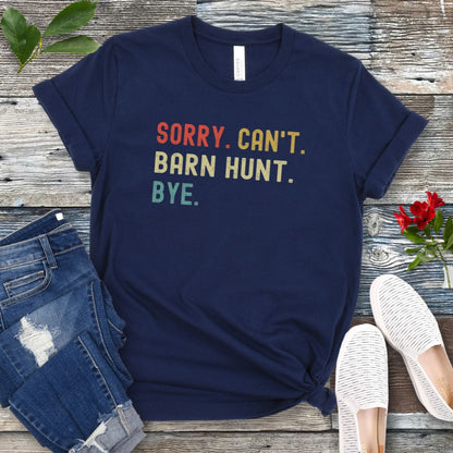 Navy blue dog sports t-shirt with colorful text 'SORRY. CAN'T. BARN HUNT. BYE.' on a wooden surface with jeans and shoes.