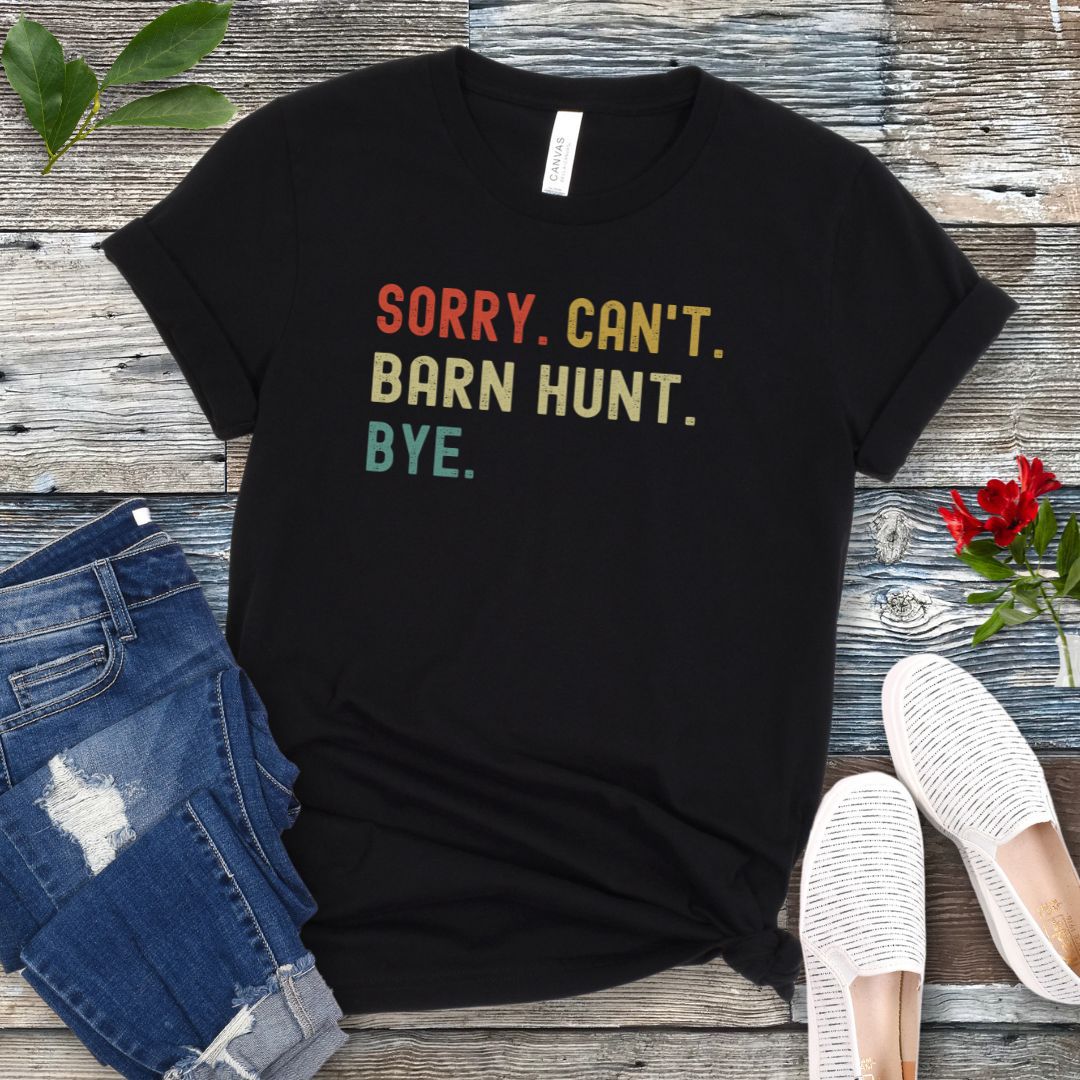 Black t-shirt with colorful text 'SORRY. CAN'T. BARN HUNT. BYE.' on a wooden surface with jeans and shoes.
