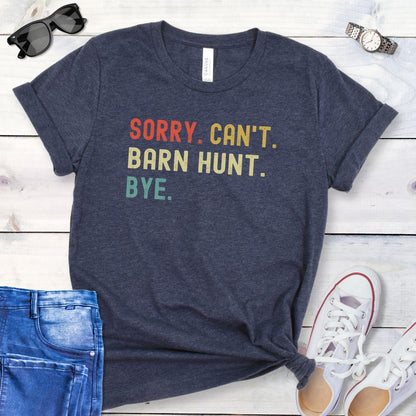 Heather navy t-shirt with colorful text 'SORRY. CAN'T. BARN HUNT. BYE.' on a wooden surface with jeans and shoes.