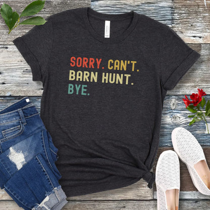 Dark grey dog sports t-shirt with colorful text 'SORRY. CAN'T. BARN HUNT. BYE.' on a wooden surface with jeans and shoes.