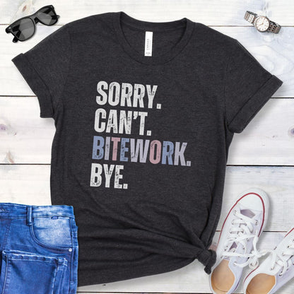 Dark grey protection dog training t-shirt with colorful, distressed text 'SORRY. CAN'T. BITEWORK. BYE.', presented on a wooden surface with jeans and shoes.