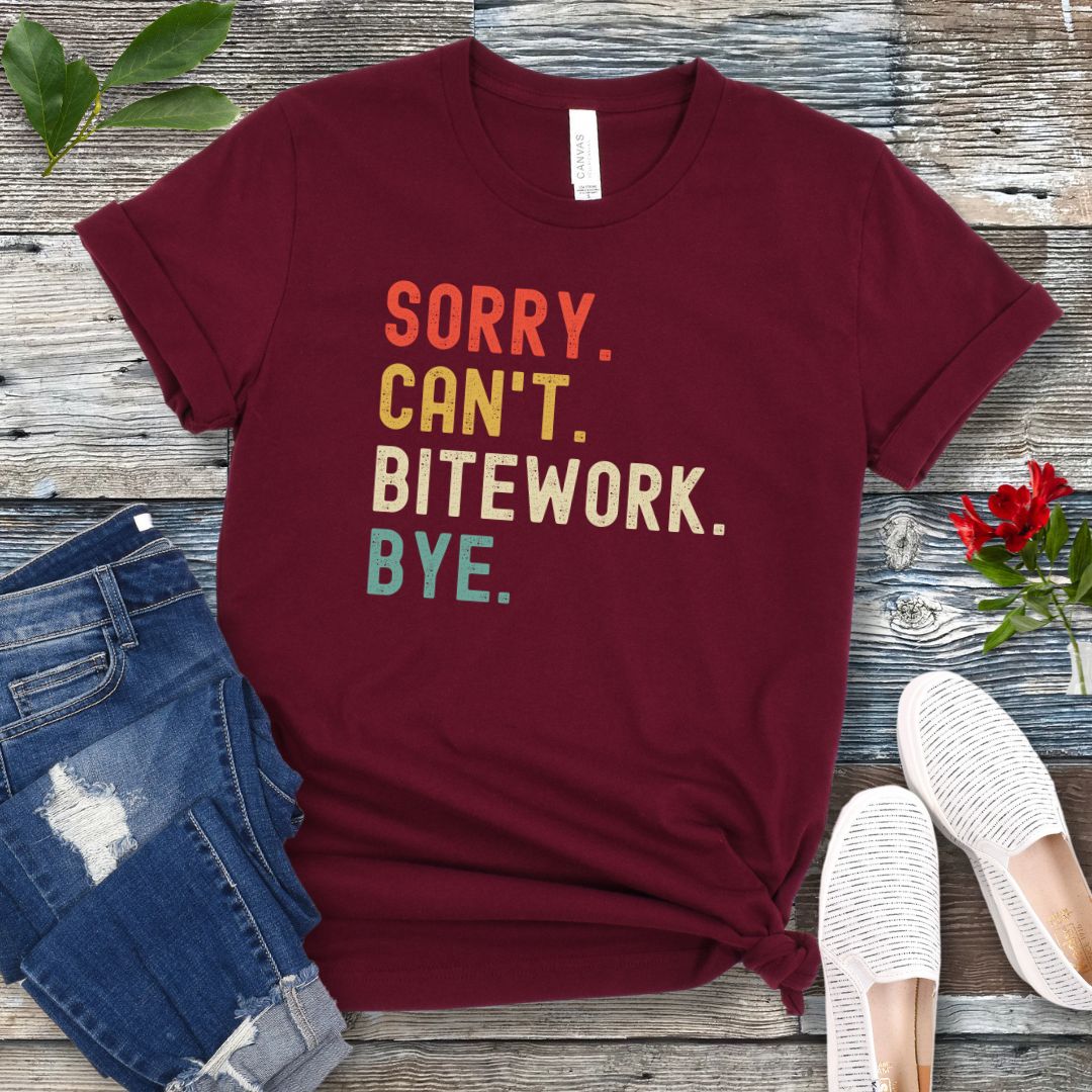 Maroon protection dog t-shirt with bold colorful text 'SORRY. CAN'T. BITEWORK. BYE.', presented on a wooden surface with jeans and shoes.