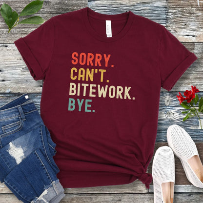 Maroon protection dog t-shirt with bold colorful text 'SORRY. CAN'T. BITEWORK. BYE.', presented on a wooden surface with jeans and shoes.