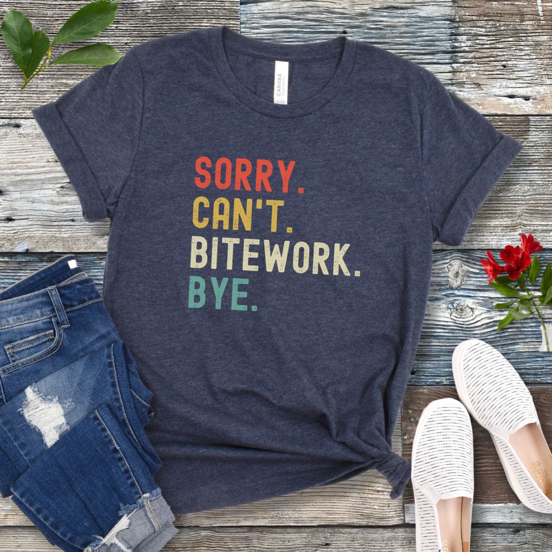 Heather navy protection dog t-shirt with bold colorful text 'SORRY. CAN'T. BITEWORK. BYE.', presented on a wooden surface with jeans and shoes.