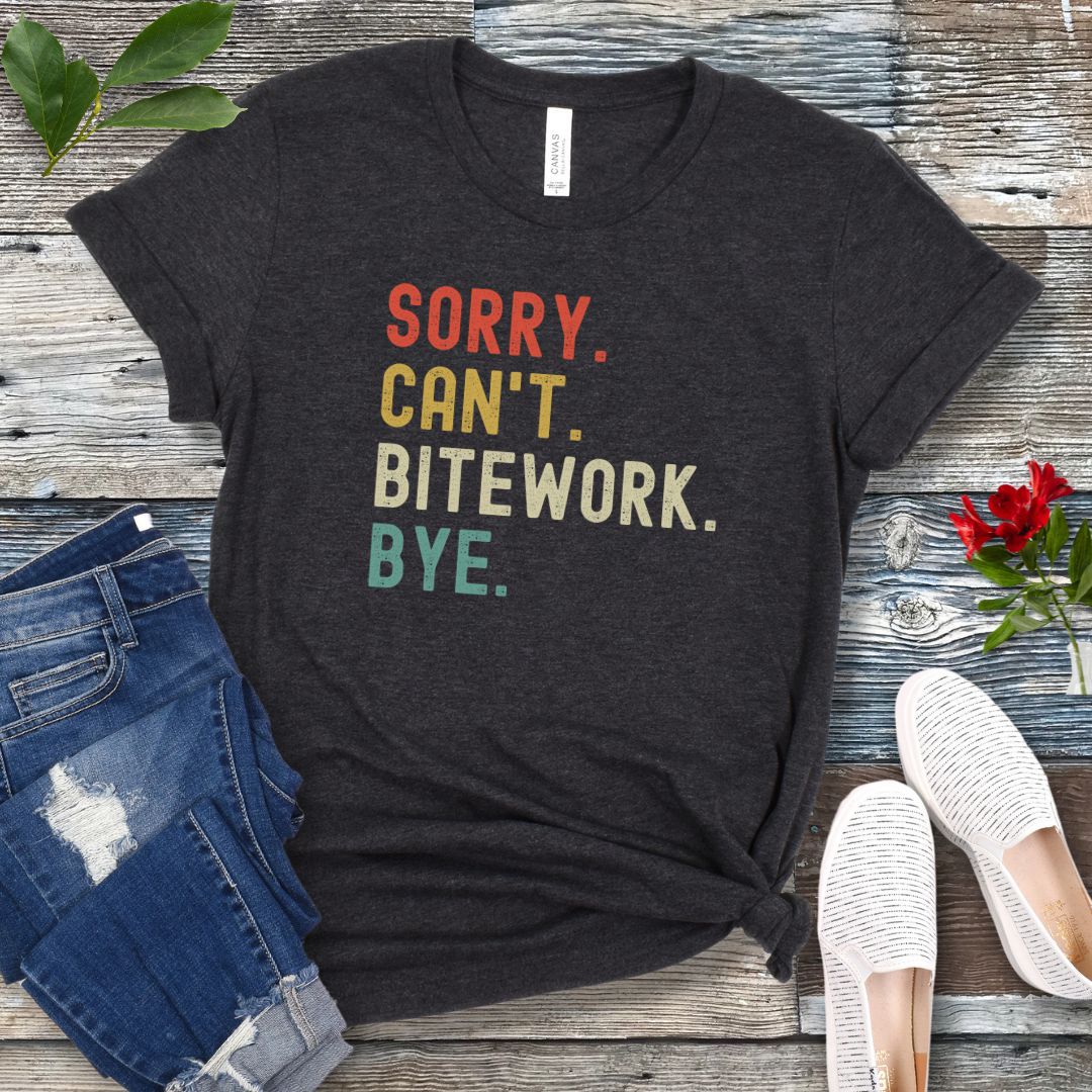 Dark grey protection dog t-shirt with bold colorful text 'SORRY. CAN'T. BITEWORK. BYE.', presented on a wooden surface with jeans and shoes.