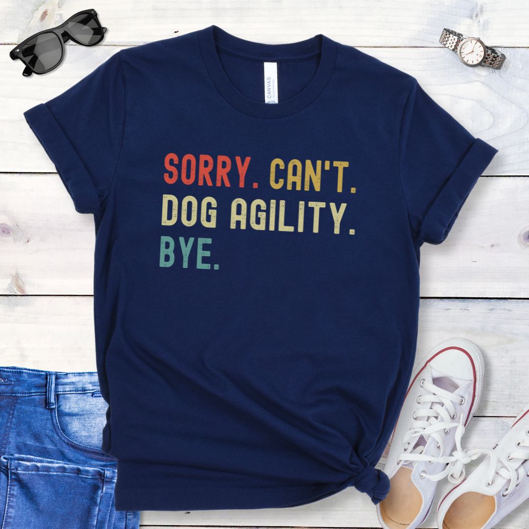 Navy t-shirt with bold, colorful 'SORRY. CAN'T. DOG AGILITY. BYE' text on a wooden surface with jeans and shoes.