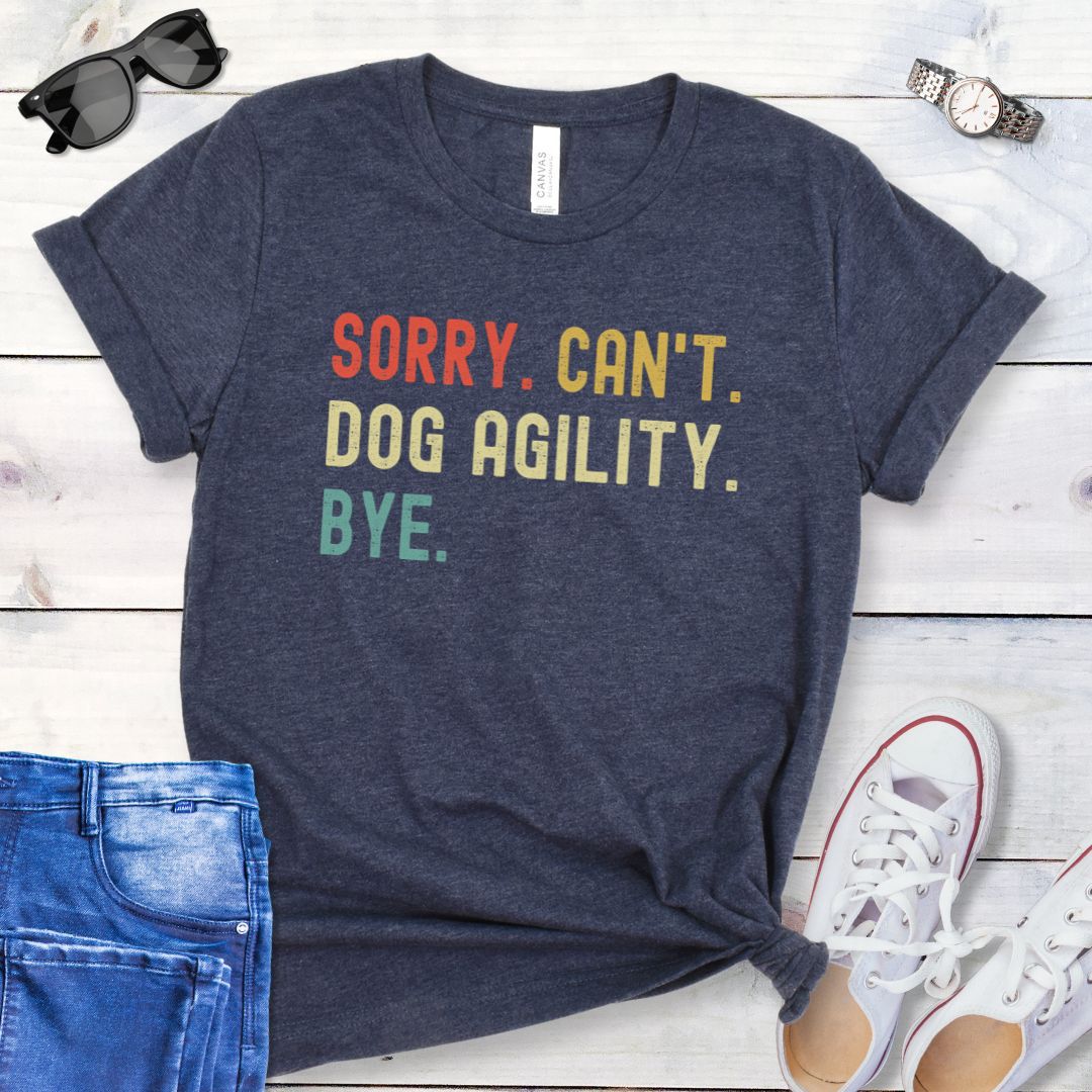 Heather navy t-shirt with bold, colorful 'SORRY. CAN'T. DOG AGILITY. BYE' text on a wooden surface with jeans and shoes.