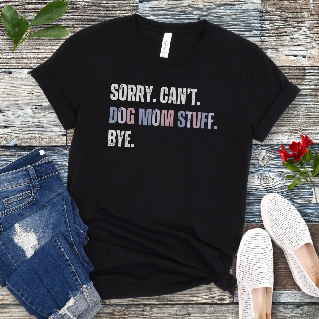 A black dog lover t-shirt with the text 'SORRY, CAN'T, DOG MOM STUFF, BYE' printed on it, displayed flat with a pair of blue jeans and white shoes, on a wooden surface with a red flower in the corner.