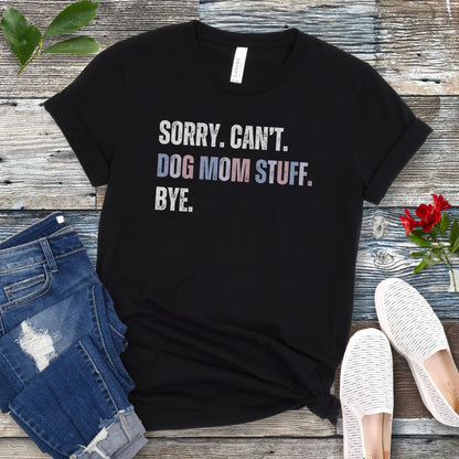A black dog lover t-shirt with the text 'SORRY, CAN'T, DOG MOM STUFF, BYE' printed on it, displayed flat with a pair of blue jeans and white shoes, on a wooden surface with a red flower in the corner.