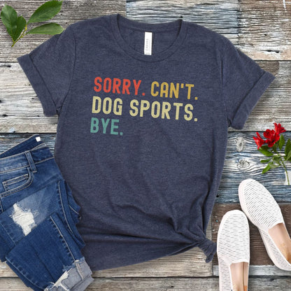 Heather navy t-shirt with the text 'SORRY, CAN'T, DOG SPORTS, BYE' in colorful letters, presented on a wooden surface accompanied by jeans and white shoes.