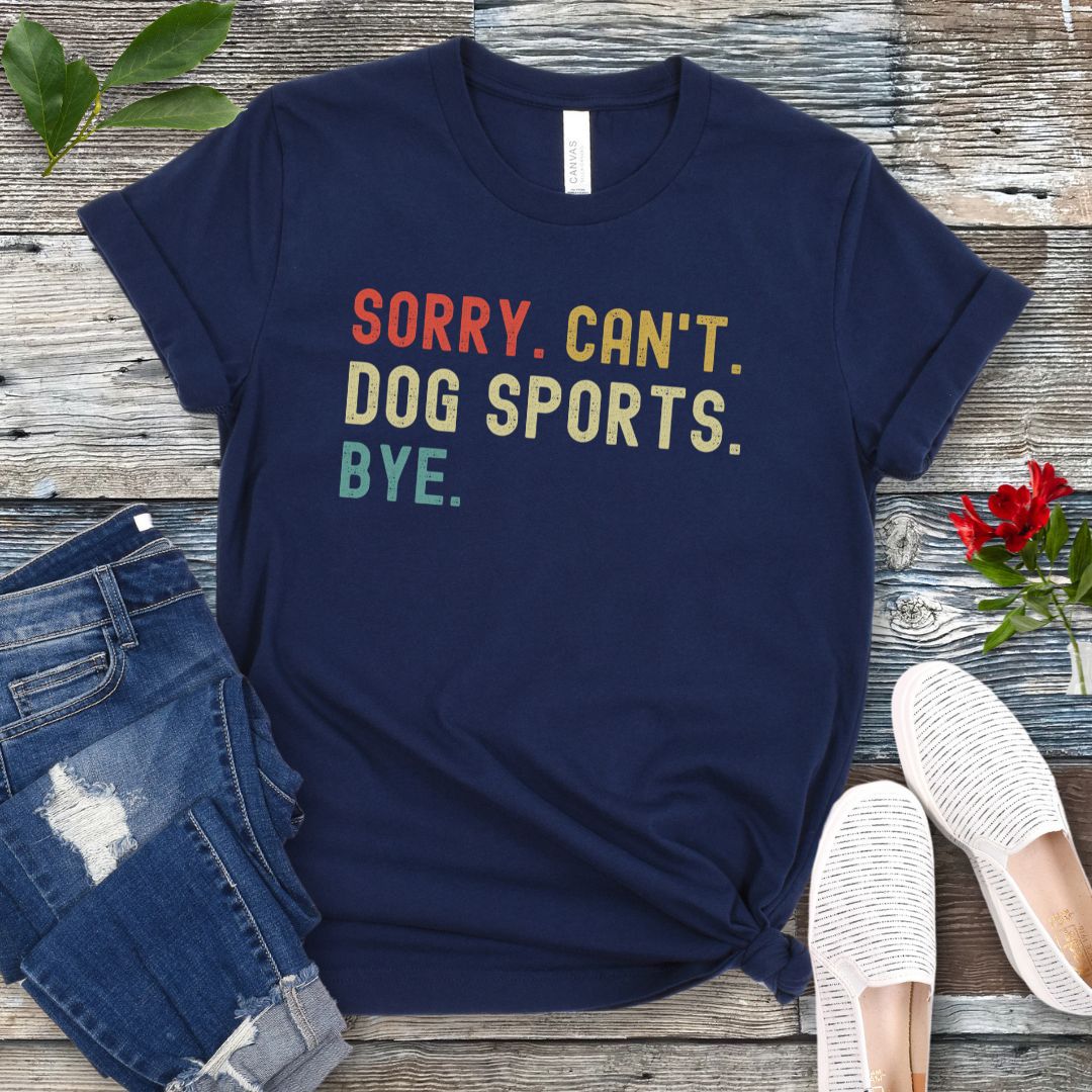 Navy blue t-shirt with the text 'SORRY, CAN'T, DOG SPORTS, BYE' in colorful letters, presented on a wooden surface accompanied by jeans and white shoes.