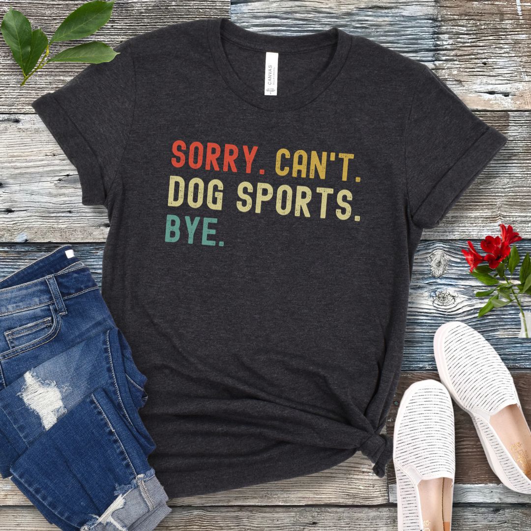 Dark grey t-shirt with the text 'SORRY, CAN'T, DOG SPORTS, BYE' in colorful letters, presented on a wooden surface accompanied by jeans and white shoes.