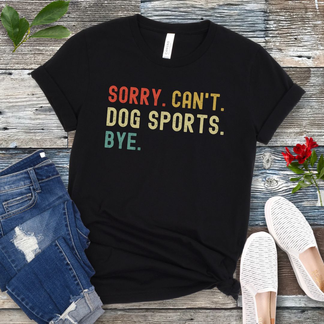 Black t-shirt with the text 'SORRY, CAN'T, DOG SPORTS, BYE' in colorful letters, presented on a wooden surface accompanied by jeans and white shoes.