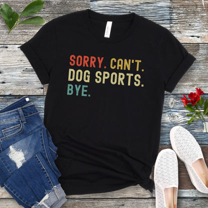 Black t-shirt with the text 'SORRY, CAN'T, DOG SPORTS, BYE' in colorful letters, presented on a wooden surface accompanied by jeans and white shoes.