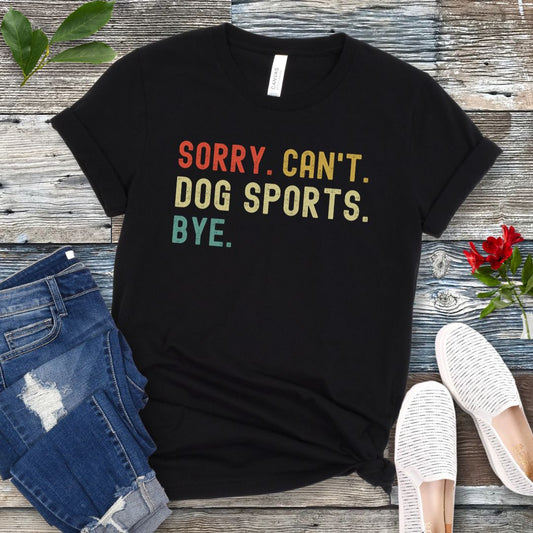 Black t-shirt with the text 'SORRY, CAN'T, DOG SPORTS, BYE' in colorful letters, presented on a wooden surface accompanied by jeans and white shoes.