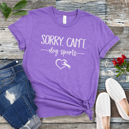 A purple T-shirt with the text 'SORRY, CAN'T dog sports' and a heart graphic with dog leaping through it under the text, displayed on a wooden background with a pair of jeans and white shoes.