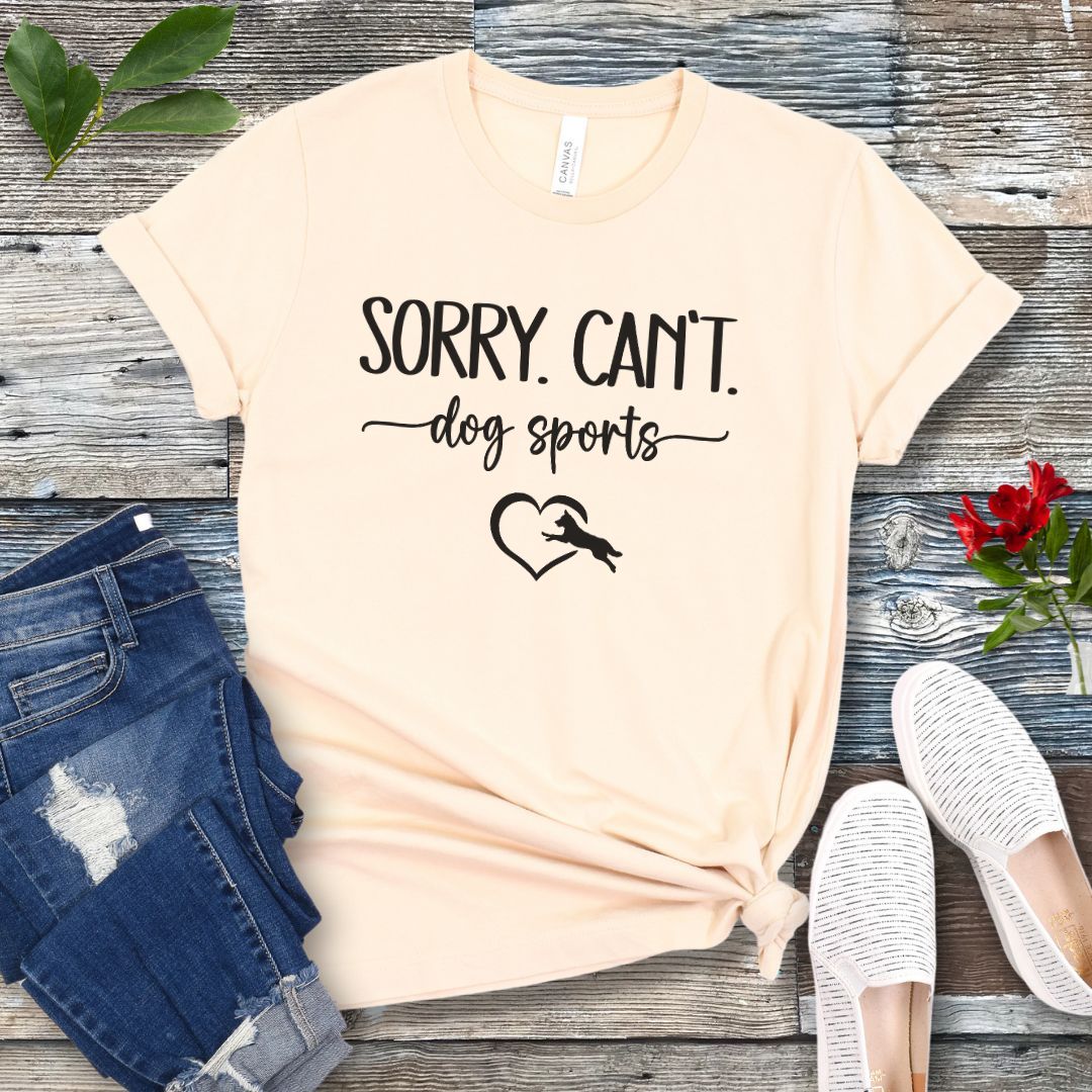 A beige T-shirt with the text 'SORRY, CAN'T dog sports' and a heart graphic with dog leaping through it under the text, displayed on a wooden background with a pair of jeans and white shoes.